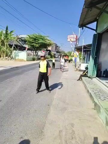 The video shows a street scene with several people walking on the right side of the road. In the foreground on the left, a person wearing a bright yellow and black vest and black pants stands facing the camera. They are holding a black object in their right hand. The person is wearing a face mask and a cap. Several other people, mostly young males, are walking away from the camera on the right side of the road, also wearing similar yellow vests. One person in the group is on a scooter. On the far right, a person is sitting on a curb, observing the scene. The environment appears to be a semi-urban or rural area with buildings and vegetation visible on the left side of the road. The sky is clear and blue, suggesting it is daytime. There are power lines running overhead.