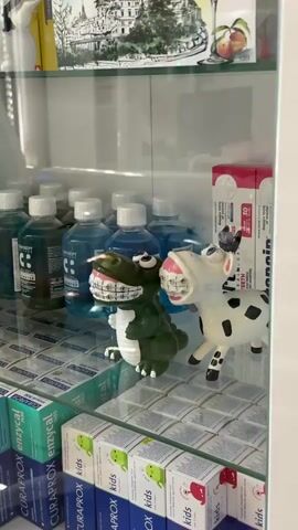 The video pans across a glass-fronted cabinet filled with dental hygiene products. On the top shelf, there are several bottles of blue and clear liquid, likely mouthwash. Towards the front of this shelf, two novelty figurines are positioned: a green alligator and a white cow, both with braces. Below this shelf, rows of small boxes are neatly stacked, labeled "CURAPROX enzycal" and "CURAPROX kids." The video then shifts to show more shelves within the cabinet, containing various bottles and boxes. To the left of the cabinet, a white printer is visible with a blue Stitch plush toy standing on it, holding a white tube that appears to be a dental product. The overall environment suggests a dental office or a retail space specializing in dental care.