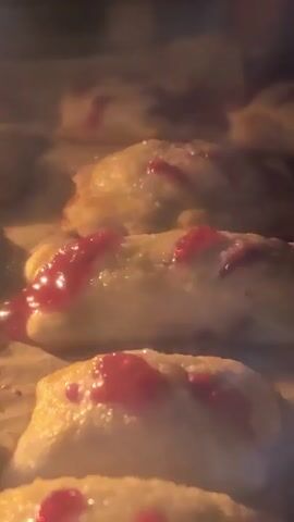 The video shows a close-up shot of several pastries with a red jam filling baking in an oven. The pastries are arranged in rows on a baking sheet lined with parchment paper. The lighting suggests the oven is on, and the pastries appear to be golden brown and slightly puffed. The red jam is visible on top of some of the pastries, and it looks glossy, indicating it is warm and possibly bubbling slightly. The overall impression is one of delicious baked goods in the process of being cooked.