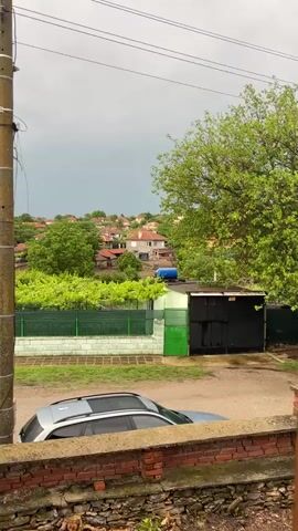 The video captures a static, slightly elevated view of a village landscape, identified as Ботево, Bulgaria, under a heavily overcast sky. In the immediate foreground, a tall, weathered concrete utility pole with multiple wires stands prominently on the left. Below the pole, partially obscured by a low brick wall, a silver car is visible, possibly an SUV.

Beyond these foreground elements, the scene reveals a collection of houses with distinctive orange or red tiled roofs, interspersed among lush green trees and dense foliage. A long, green metal fence runs across the mid-ground, behind which vibrant green leafy plants, potentially grapevines, are visible. One house in the background features a blue cylindrical water tank on its roof. Further beyond the village houses and trees, a wide expanse of open, flat land or fields stretches towards the horizon under the somber, gray clouds.

The weather appears cloudy, suggesting a cool or potentially rainy day, and the lighting is diffused. No people are visible in the scene, and there is no discernible activity. No visible text appears in the video. The overall impression is of a quiet, rural village setting.