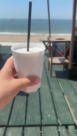 The video captures a bright, sunny day at a beachfront location in Nέα Χηλή, Greece. It begins with a close-up shot of a hand holding a white, faceted plastic cup containing a milky-white drink with a black straw. The hand has light pink nail polish. The camera then pans to reveal a wooden deck, appearing to be part of a beach bar or cafe, with a greenish hue. Beyond the deck is a sandy beach leading to the calm, blue ocean. In the distance, a small boat is visible on the water under a clear blue sky. The deck is furnished with wooden tables and chairs, and shaded by structures with fringed awnings or umbrellas. The scene evokes a relaxed, summery atmosphere by the sea. No text is visible.
