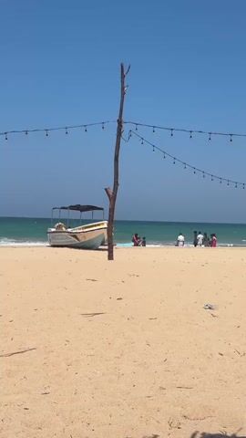 A wide shot of a sandy beach on a bright, sunny day under a clear blue sky. In the foreground, the sand is smooth and light brown. A thin, vertical wooden pole stands in the middle of the frame, from which strings of small, round lights are strung horizontally across the sky. To the left, a light-colored motorboat with a dark canopy is partially visible, resting on the sand near the water's edge. In the background, the turquoise ocean waves gently break on the shore. A group of approximately five people are gathered near the water, some appearing to be sitting or standing. The overall scene evokes a peaceful beach atmosphere.