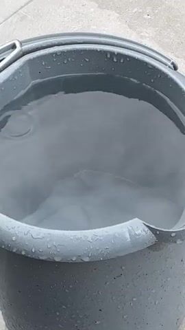 A grey bucket is partially filled with water. Water drops are falling from above, creating ripples and splashes on the surface of the water in the bucket. The bucket appears to be made of plastic and has a metal handle. The surface surrounding the bucket is made of grey paving stones.