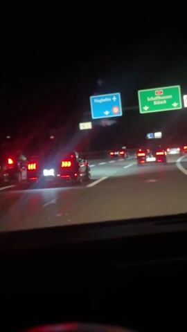 The video shows a view from inside a car, driving on a highway at night. Several cars are visible ahead, including multiple Ford Mustangs, identifiable by their distinct three-bar tail lights. The road has white lane markings. Overhead highway signs indicate directions to "Flughafen" (Airport) and "Schaffhausen" and "Bulach". The overall environment appears dark.