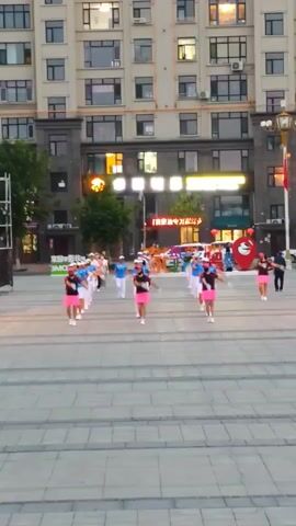 A group of women in pink skirts and black tops, some with white gloves, are performing a coordinated dance in an outdoor plaza. They are arranged in rows and appear to be moving in unison. Behind them is a large, modern building with multiple floors and windows. A few other people are visible in the background, including a person in a white shirt and black pants, and a person in a blue shirt. There are also some signs and decorations on the building, including a red heart-shaped object. The ground is paved with large, light-colored stones. The lighting suggests it is late afternoon or early evening.
