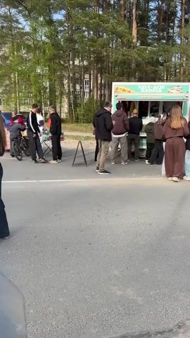 A mobile food truck is parked on the side of a road with a group of people queued up in front of it. The truck is decorated with images of food. The text on the side appears to say "Хот-дог завод," which translates to "Hot dog factory" in English. People in the line are wearing casual clothes and appear to be waiting to purchase food. There are trees in the background. The road is asphalt and there is a small sandwich board to the left of the truck.