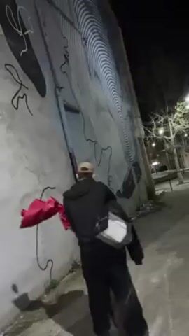 The video shows two people, presumably a couple, on an urban sidewalk at night. One person, wearing all black with a beige cap and a white over-the-shoulder bag, holds a bouquet of red flowers and an article of clothing. They are embracing the other person, who is also in dark clothing. The scene takes place alongside a wall with a large, black and white mural. Streetlights and bare tree branches are visible in the background. The camera is hand-held and slightly shaky. The person filming the scene says, "So cute. So cute."