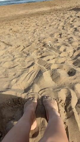 The video begins with a close-up shot of a person's feet partially buried in the sand on a beach. The camera then slowly pans upwards, revealing more of the sandy beach with some faint footprints. In the distance, the ocean can be seen with gentle waves and a clear blue sky. A few yellow buoys are visible in the water. The video concludes with a shot of the beach and the ocean, with a pair of flip-flops lying on the sand in the foreground.