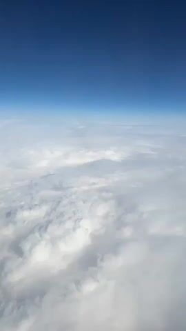The view from an airplane window shows a sea of white clouds stretching to the horizon, where they meet a clear, bright blue sky. The clouds are dense and puffy, with varying shades of white and gray. The sky is a uniform, deep blue color. The scene is bright and airy.