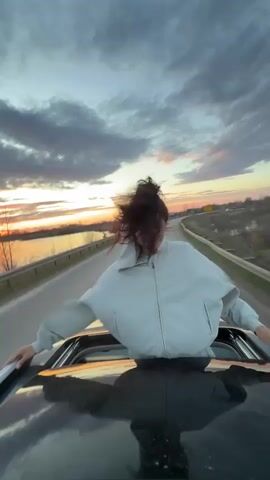 A young woman is captured standing joyfully through the sunroof of a dark-colored car, likely moving along a road in Dubnik, Russia. Her dark hair is swept back and wildly blown by the wind, framing a wide, happy smile. She is wearing a light grey or white zip-up hooded jacket and dark pants, with her arms resting on the car's roof, hands gripping the edges of the sunroof opening.

The scene unfolds during either sunset or sunrise, with a vibrant orange and yellow glow visible on the horizon, casting a warm light on the dramatic, cloudy sky. The road runs parallel to a body of water, possibly a river or lake, on the left side of the frame. Beyond the water, a flat landscape with distant trees is visible. The right side of the road features a more open, possibly grassy or barren area. The car's reflective roof shows a blurred reflection of the sky and surroundings.

The overall atmosphere conveys a sense of freedom, exhilaration, and happiness, with the woman embracing the moment and the open air. There is no visible text in the frame.