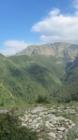 This video showcases a scenic mountain landscape in Kalofer, Bulgaria, under a partly cloudy sky. The scene transitions from a wide view of lush green valleys and rugged mountain peaks to a closer shot of a trail marker post. This post, painted with alternating yellow and black stripes, stands firmly on a rock amidst a rocky terrain with sparse vegetation. In the final frames, a person with a backpack is seen walking away from the camera along a mountain path, hinting at a hiking or trekking activity. The overall mood is one of adventure and appreciation for nature's grandeur. The time of day appears to be daytime, with ample natural light illuminating the landscape. No visible text is present in the video.
