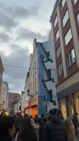 The video shows a street in Brussels, Belgium, on a cloudy day. The street is lined with tall buildings, including one with a mural of characters climbing a set of blue and white stairs. A crowd of people is walking down the street, and there are shops on either side of the street. A street sign with an arrow is visible.