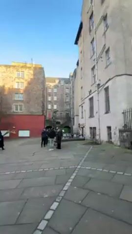 The video shows a stone-paved square surrounded by tall, old buildings under a bright blue sky. The buildings are varied in color, ranging from beige to grey. Some have red sections, while others have numerous windows. Several groups of people are visible, some standing and some walking. A lamppost stands in the square. A building in the foreground features a doorway and a sign.