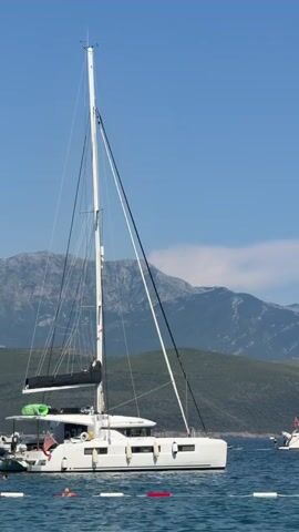 The video shows a wide shot of a bay with a clear blue sky and distant mountains. In the foreground, a white catamaran with a tall mast is anchored in the water. To the right of the catamaran, a white motor yacht is also anchored. The water is calm and there are a few buoys visible in the foreground. The overall impression is one of a peaceful and scenic maritime setting.