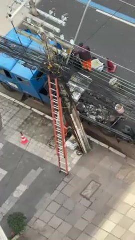 The video shows a blue utility truck parked next to a utility pole, with a person working on the complex arrangement of overhead wires. The truck bed is filled with debris. An orange ladder is leaning against the pole, providing access to the wires. A traffic cone is visible on the sidewalk. The scene is filmed from an elevated perspective, looking down onto the truck, pole, and adjacent street and sidewalk. The sidewalk is made of gray interlocking bricks.