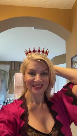A woman with blonde hair and red lipstick is wearing a crown and a pink robe with black lace. She is smiling and touching her hair. The background appears to be a hallway with an arched doorway and a light fixture on the ceiling. There are framed pictures on the wall and a shower door visible in the background.