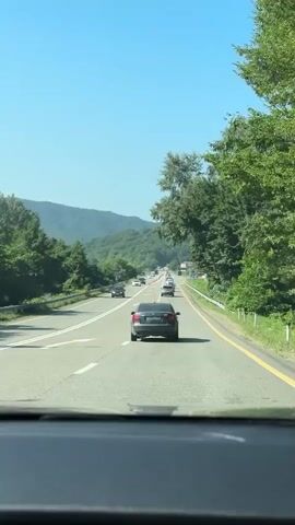 The video is filmed from inside a vehicle, looking out through the windshield. The camera is focused on the road ahead, which is a multi-lane highway or major road. Several cars are traveling in the same direction. The road is surrounded by lush green trees and rolling hills under a clear blue sky. The perspective suggests the vehicle is in motion, likely driving on this road. The interior of the car, specifically the dashboard area, is visible in the foreground, slightly out of focus. The overall atmosphere is that of a scenic drive during the daytime.