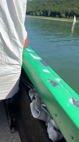 The video shows a scenic view of a lake with sailboats. The lake water is a deep green color, and the surface is rippling. In the background, a lush green hillside covered in trees slopes down to the water's edge. The sky is a clear blue with a few wispy white clouds. Several white sailboats are visible on the water, appearing to be engaged in sailing. Their sails are white and triangular. One boat in the foreground on the right side is leaving a white wake. The scene suggests a sunny day, possibly with a gentle breeze, ideal for sailing. There are no people clearly visible on the boats.