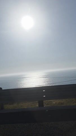 The video shows a bright sun over the ocean. The camera is looking out at the ocean from behind a guardrail. There is some vegetation and a road in the foreground, in front of the guardrail.