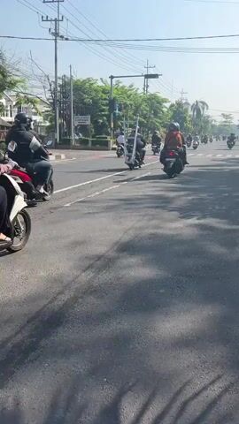 The video shows a group of motorcyclists riding down a street. The motorcyclists are wearing helmets and riding on scooters. The street is paved and has traffic lights and power lines. There are trees and buildings on the side of the street. The sun is shining and the sky is clear. The video is taken from a low angle, looking up at the motorcyclists. The video is in color and the sound is of the motorcycles.