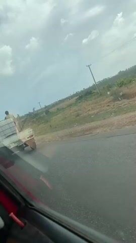 A white mini-truck, possibly a van with the top cut off, is driving on an asphalt road. A large, caged rectangular container, possibly a water tank, is secured to the bed of the truck. A person wearing a dark shirt and hat is riding on top of the container. Another person is standing near the truck's driver's side. The license plate of the truck is partially visible. The surrounding environment features a green field, a utility pole, and a cloudy sky. The footage appears to be taken from a moving vehicle.