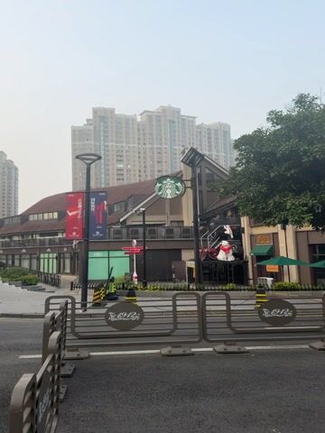 The scene shows an exterior shot of a Starbucks storefront in a commercial district. The Starbucks logo is prominently displayed on the building. In front of the building, there are gray metal barricades with circular designs. Behind the Starbucks building are several tall apartment buildings. There are flags and banners on the building. The sky appears overcast. The environment suggests an urban setting. There are trees on the right side of the frame. No people are clearly visible.