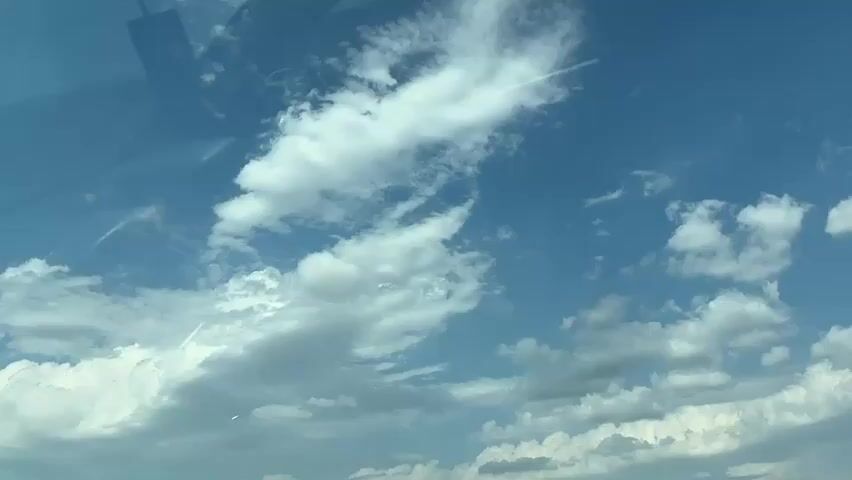 The video shows a view of a blue sky with white clouds. There are faint reflections visible on the left side of the screen, suggesting the footage was captured through a glass surface, possibly a car windshield. A few contrails are visible in the sky, indicating the presence of aircraft. The clouds are varied in density, with some appearing wispy and others more solid. The overall impression is of a sunny day with scattered cloud cover.