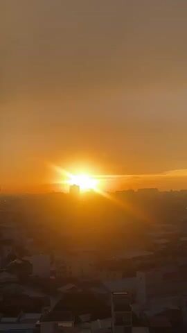 This is a vertical video of a sunset over a city. The sun is a bright, glowing orb in the center of the frame, casting long rays of light across the sky. The sky is a gradient of orange and yellow hues. In the foreground, silhouetted buildings and rooftops are visible, suggesting an urban environment. The light is warm and soft, creating a peaceful and serene atmosphere. The time of day is clearly sunset, indicated by the low position of the sun and the warm, golden light. There are no people or specific objects clearly identifiable in the foreground due to the silhouette effect. The overall mood is tranquil and visually striking, highlighting the beauty of the natural phenomenon against the backdrop of the city. No text is visible in the video. The context provided indicates the location is Khan Mean Chey, Cambodia.