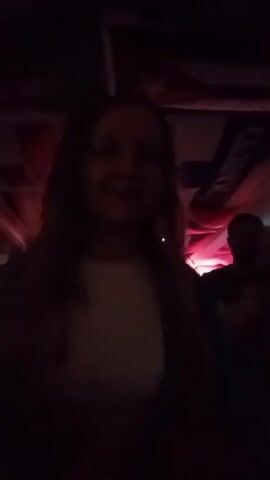 A dark, dimly lit room with red lighting accents. In the foreground, a woman with long, dark hair is silhouetted against a white shirt and dark jacket. Her arms are raised above her head. To her right, another person is partially visible, also in shadow. In the background, a man in a light-colored shirt can be seen. Above, there appears to be a ceiling structure with what could be flags or banners draped. The atmosphere suggests a dimly lit venue, possibly a club or concert hall.