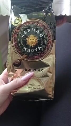 The video shows a hand with long, painted fingernails holding a bag of coffee. The coffee bag is gold and black with a circular logo featuring the sun and the words "ЧЕРНАЯ КАРТА" in Russian. Text below the logo reads "В ЗЕРНАХ" and "СРЕДНЯЯ ОБЖАРКА," indicating whole beans and medium roast. The bag is slightly crinkled. The background is out of focus, showing what appears to be furniture and possibly a curtain. The lighting is moderate.
