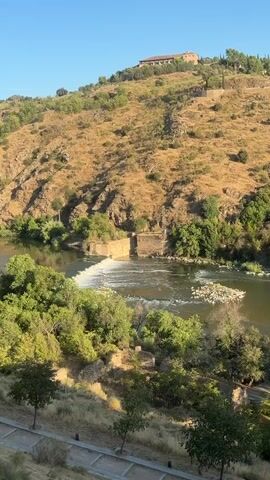 The video shows a wide, slow-moving river flowing through a valley. The river has a small waterfall or dam in the middle, causing white water to churn. The banks of the river are lined with lush green trees, and further back, a steep, dry, grassy hill rises. On top of the hill, a large building with a reddish-brown roof is visible. The sky is clear and blue, indicating a sunny day. In the foreground, a pathway or road runs along the bottom of the frame, with a few small trees planted nearby. The overall impression is a serene and natural landscape.