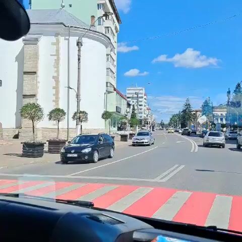 The video shows a view from inside a vehicle driving down a street. The street has multiple lanes and is lined with buildings on both sides. There are several cars on the road, moving in the same direction as the camera. The sky is blue with some clouds. On the left side of the road, there are trees in planters and a building with a curved white facade. On the right side of the road, there are more buildings, trees, and power lines. Traffic signs and pedestrian crossing markings are visible on the road. The overall scene depicts a typical urban environment on a sunny day.