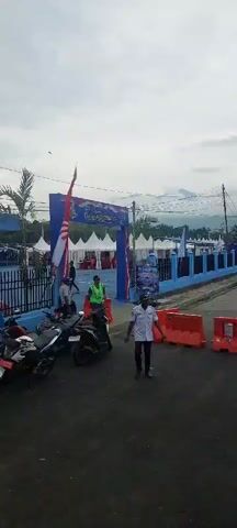 The video shows an outdoor event with a large blue archway that says "Festival Danau Sentani XV." The Indonesian flag is displayed prominently on the left side of the archway. Several white tents are set up behind the archway, with people visible inside some of them. Two men in uniform are standing near the entrance, one in a white shirt and the other in a green vest. A motorcycle is parked on the left. Orange barriers are placed on the ground. The sky is overcast, and mountains are visible in the background. There are also trees and power lines in the scene. The overall impression is of an organized outdoor festival or gathering.