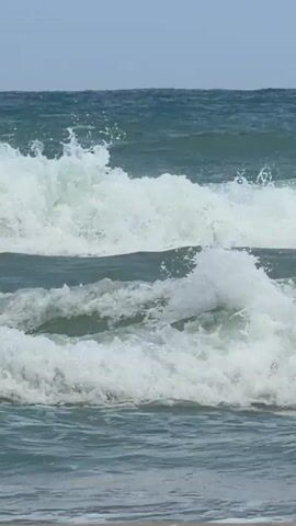 A video of ocean waves crashing on a sandy beach. The waves are moderate in size with white foam crests. The water is a mix of blue and green hues. The sky is overcast and the horizon is visible. In the middle ground, a green rope is visible floating on the water. The camera is static and focused on the waves.