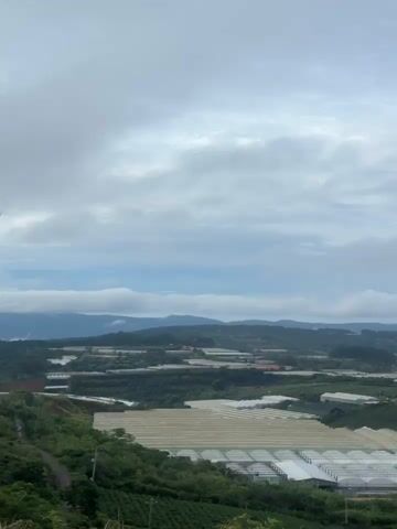 The video presents a scenic view of a mountainous landscape under a cloudy sky. Initially, a wide expanse of greenhouses dominates the foreground, gradually revealing a broader view of a town nestled amidst the hills. The landscape features lush greenery and distant mountain ranges. As the video progresses, a prominent wind turbine comes into view, positioned atop a hill overlooking the town. The turbine is white with three blades, standing tall against the backdrop of the town and distant mountains. The camera pans slightly, showcasing another wind turbine in the distance. The overall scene suggests a blend of agricultural land, residential areas, and renewable energy infrastructure in a rural, mountainous setting.
