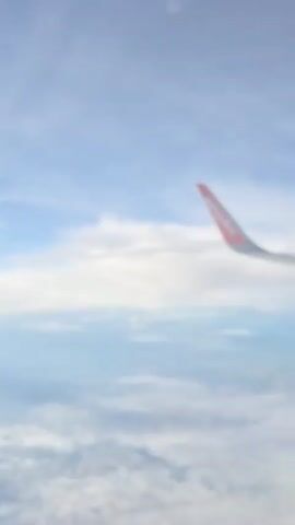 The video is a point-of-view shot from an airplane window. The EasyJet logo is visible on the wingtip in the upper right quadrant of the frame. Below the wing, a blanket of clouds stretches across the horizon, with hints of land visible through breaks in the clouds. The sky is a pale blue with scattered white clouds. The perspective suggests the airplane is in flight, with the wing pointing upwards and slightly to the right.