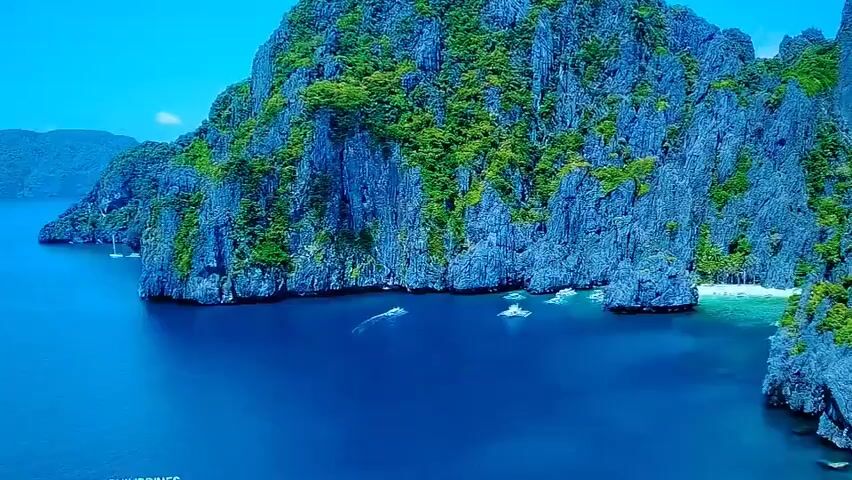 An aerial drone shot shows a clear, blue ocean meeting a rocky, jungle-covered island. Several small boats are visible on the water near the shore. The sky is a bright, clear blue.