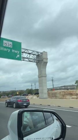 The video shows a view from inside a car, looking out the passenger side window. The car is on a road with traffic. There is a grey sedan in the lane to the left, and construction barrels on the median. Across the road, there are some businesses, including one that appears to be a coffee shop, as well as power lines and trees. The sky is overcast. The side mirror of the car is visible, showing a reflection of the interior and the road behind.  The text "OBJECTS IN MIRROR ARE CLOSER THAN THEY APPEAR" is visible on the mirror.