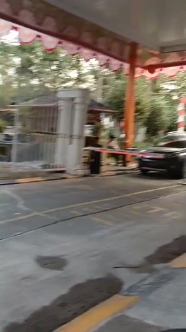 A dark-colored SUV is positioned at an exit gate. A person wearing a light-colored shirt and dark pants is seated behind a white booth. A boom barrier, painted green, is in the process of being lifted. The gate structure is adorned with red and white decorative trim. The ground is paved, with yellow markings visible. Trees and foliage can be seen in the background.