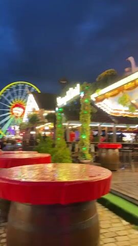 The video shows a bustling outdoor fair at dusk. In the foreground, there are round tables covered with red cloths, supported by barrel-like bases. Behind these tables, a wooden deck area is visible with more seating and a bar or food stall decorated with lights and greenery. A sign with German text and the number 10.50 is on the left. In the mid-ground and background, a large Ferris wheel illuminated with colorful lights dominates the scene. Numerous people are walking around, some gathered in groups. Other fair attractions with bright lights are also present in the distance. The sky is dark blue with some clouds, indicating evening or night. The overall atmosphere appears lively and festive.