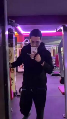 A man wearing glasses, a black hoodie, and black pants holds a phone and takes a selfie in a gym with purple and red lighting. The gym contains various weightlifting machines and equipment. The camera then pans across the gym interior showing the weightlifting machines and other equipment.