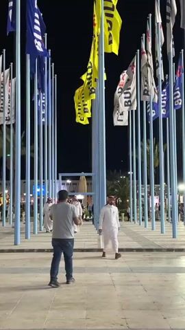 The video shows a plaza at night filled with tall poles displaying flags. The flags are a mix of blue and yellow, with white flags also visible. Many of the flags have the word "BUILD" printed on them. The ground is paved with large square tiles. Several people are walking through the plaza. In the foreground, a man in a grey shirt and blue jeans walks away from the camera. In the background, several men in traditional white thobes are walking towards the camera. The lighting suggests it is nighttime, with artificial lights illuminating the area. Palm trees are visible in the background, indicating a warm climate. The overall atmosphere is festive and public.