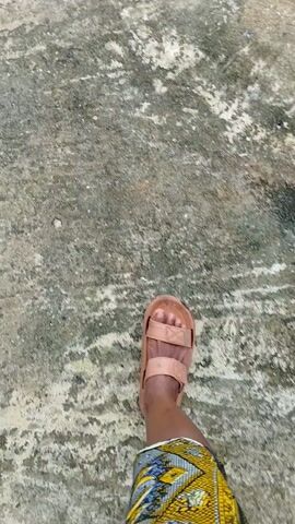 A first-person perspective video shows a person's legs and feet walking on a rough, textured surface, possibly concrete or pavement, with a slightly wet or stained appearance. The person is wearing a light brown, strappy sandal on their right foot. They are also wearing a skirt or dress with a vibrant, patterned fabric featuring yellow, blue, and white designs. The camera movement suggests the person is walking forward, and the focus is primarily on the lower body and the ground.