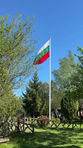 The video shows a Bulgarian flag waving in the wind on a sunny day. The flag is white, green, and red, and it is mounted on a tall, white flagpole.  The flagpole is surrounded by trees, grass, and a wooden fence. In the background, a person wearing a backpack can be seen walking through the park. The sky is clear and blue.