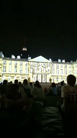 The video shows a building illuminated with a light show at night. A crowd of people are sitting on the ground in front of the building, watching the display. The building is large and appears to be made of light-colored stone. It has many windows and architectural details, which are highlighted by the light show. The light show consists of various patterns and shapes of light projected onto the building, including geometric shapes and what appear to be abstract representations of stars or confetti. The sky is dark, indicating that it is nighttime. The people in the foreground are mostly silhouetted against the light from the building. They are sitting on blankets or directly on the ground, and some are looking up at the building, while others are facing away. The overall atmosphere is one of a public event or performance.