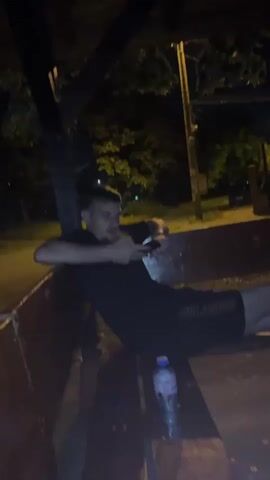 A medium shot shows a young man with short hair and a goatee sitting in a dark, outdoor setting at night. He is wearing a black t-shirt and black shorts. He is leaning back on a wooden structure, holding a smartphone in his hands and looking at it. A plastic water bottle with a blue label is visible on the wooden surface in front of him. Trees with foliage are visible in the background, illuminated by an unseen light source. A wooden post is visible on the left side of the frame, and a metal pole with lights is visible in the background.