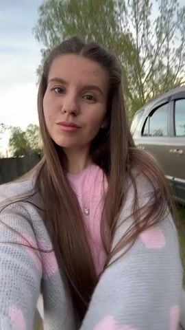 A woman with long brown hair and a light pink shirt and grey sweater is recording a selfie video outside. She is standing on a grassy area with a dirt path. A silver vehicle is parked behind her. A fence, trees, and a shed are also visible in the background. She looks directly at the camera and slowly pans the camera around the area.