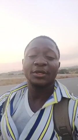 A man with dark skin and a striped shirt is recording a selfie video. He has a backpack on. He is standing on a paved road with light-colored sky in the background. In the distance, there is a vehicle. The man is making hand gestures and the audio contains a very loud roaring sound.