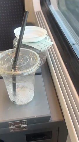 A still shot of a train tray table is shown, positioned next to a window. On the tray, there are two clear plastic cups with lids and black straws. The cup in the foreground is half-full of ice and a clear liquid, with condensation on the outside. The second cup, behind it, is mostly obscured by the first but appears to contain a dark liquid. A piece of paper or receipt is visible between the two cups. The tray itself has a metal hinge and what looks like an electrical outlet underneath. The window shows a blurry outdoor scene with what appears to be vertical blinds. The overall setting suggests a journey on a train.