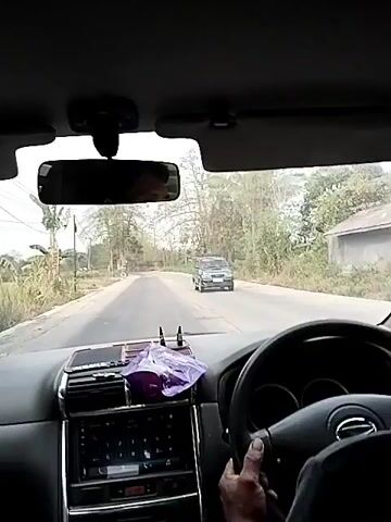 The video shows the interior of a car from the driver's perspective. The dashboard and steering wheel are visible, along with the road ahead. Another car, a green SUV, is driving in the opposite direction on the road. The scene appears to be a rural or suburban area with trees and vegetation visible on the sides of the road. The lighting suggests it is daytime.